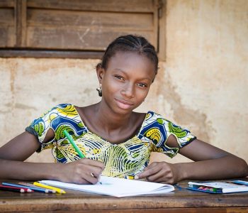 Portrait of an African ethnicity schoolgirl (age 13) in an educational environment in Bamako, Mali learning her lesson outdoors sitting on a desk and slightly smiling.